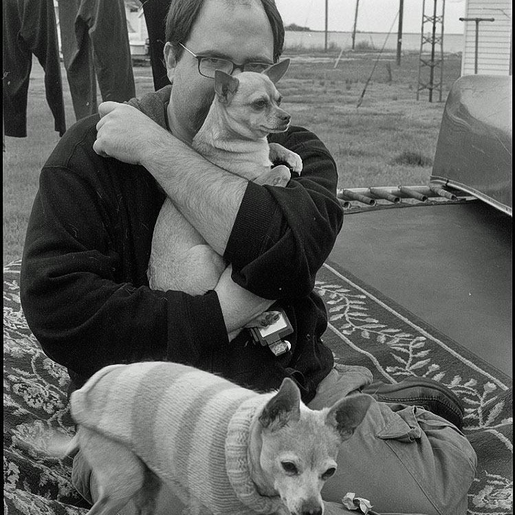 Black and white photograph of man seated on ground with two small dogs
