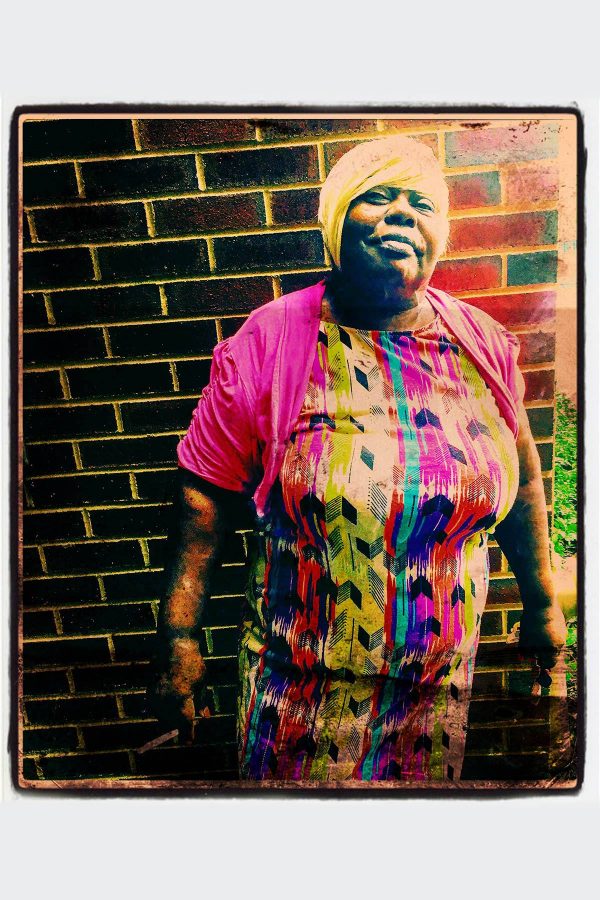 Color photograph of woman wearing bright colors, standing in front of a brick wall