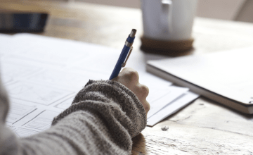 Close-up photograph of a woman's hand writing on some papers. A notebook and coffee mug litter the table beside her.